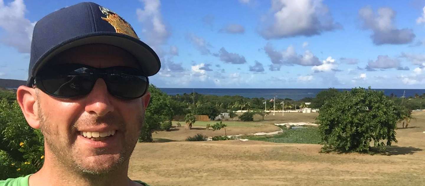 This image features Michael Barnett wearing a cap and sunglasses smiling for a photo, with the disc golf course called The Reef in the background.